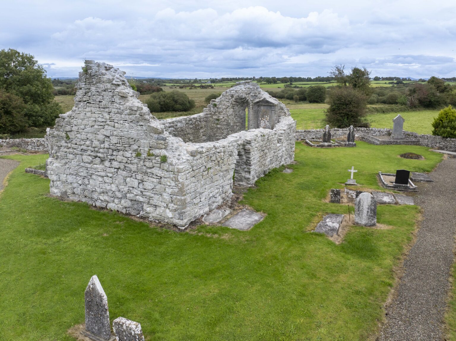 Killoo Church structure in a grassy graveyard with several gravestones, set in a rural landscape under a cloudy sky.