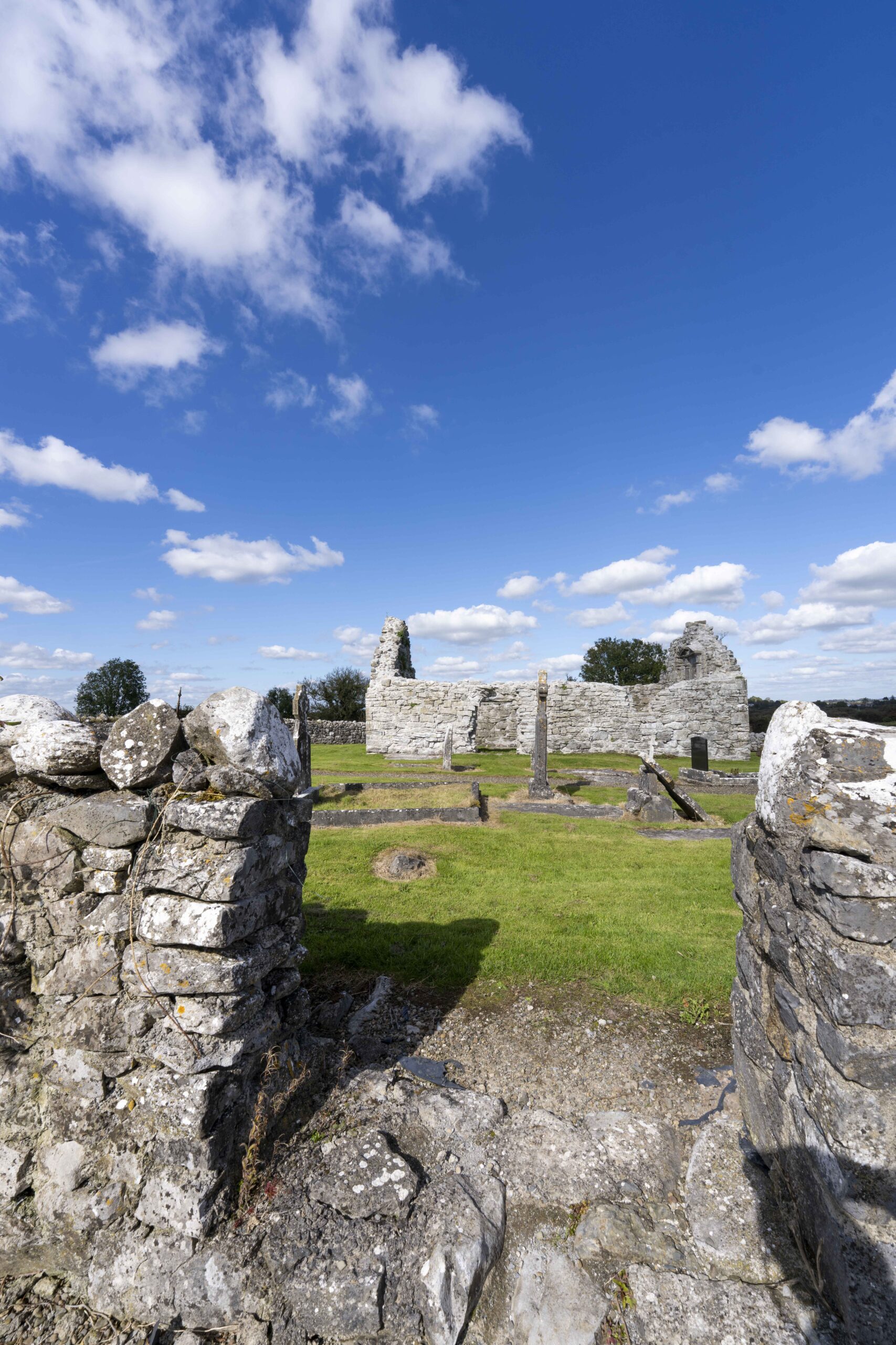 View of Killoo Church ruins with low walls surrounding a grassy area and standing stone crosses under a bright blue sky with scattered white clouds.