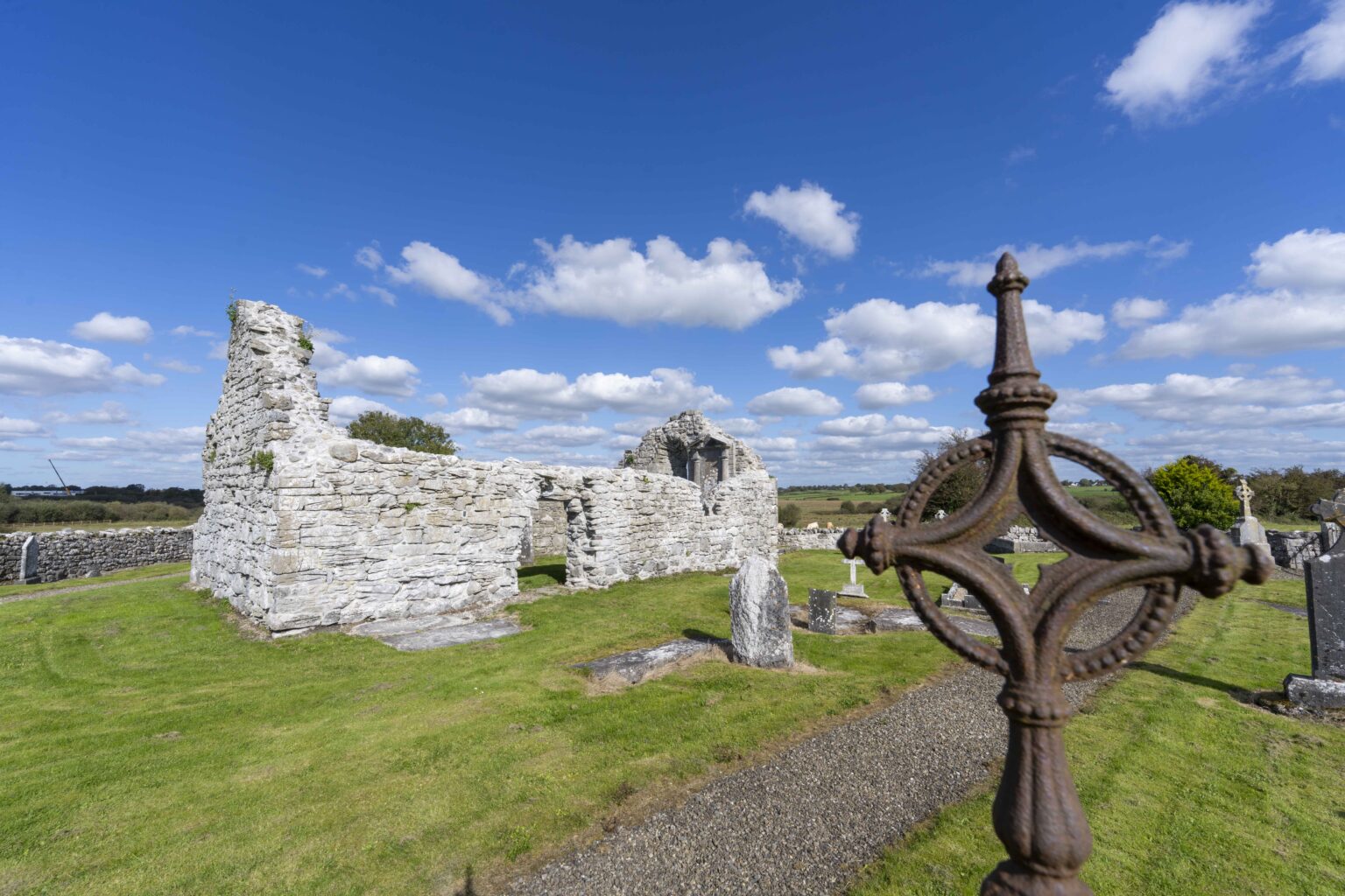 Ruins of Killoo Church in a graveyard with green grass and scattered gravestones, under a blue sky with white clouds, and a close-up of a rusty iron cross in the foreground.