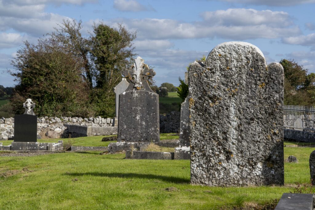 A grassy cemetery with several old, weathered headstones and crosses under a partly cloudy blue sky, with trees and a stone wall in the background.