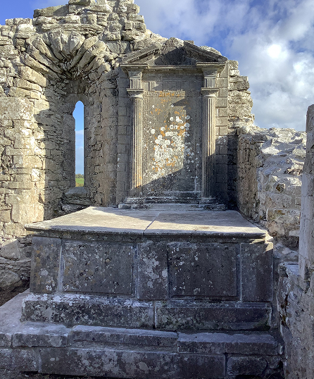 Ancient stone altar structure set within partially ruined stone walls, featuring decorative carvings on the back panel, under a partly cloudy sky.