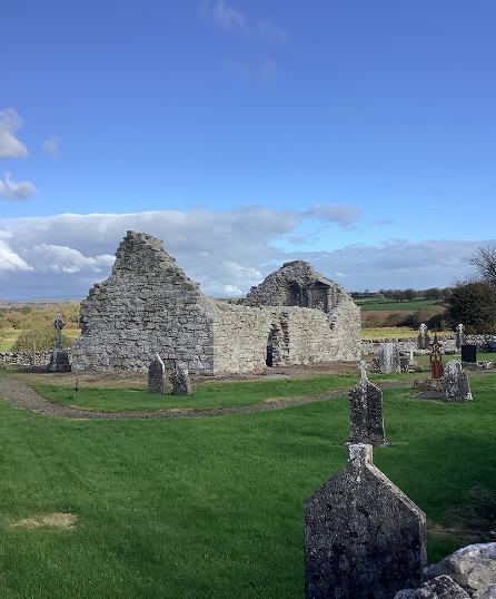 Ruins of an old stone building surrounded by a graveyard with weathered tombstones, set against a backdrop of green fields and a partly cloudy blue sky.
