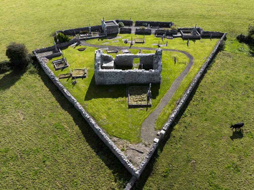 Aerial view of a triangular stone enclosure with a small stone ruin and several graves inside, set in a grassy field with a cow grazing nearby.