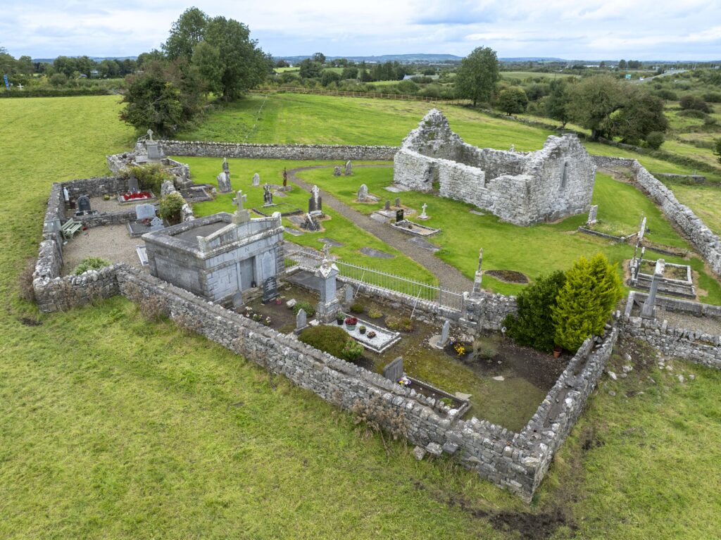 Aerial view of an old stone-walled cemetery and church ruins situated in a green, grassy rural landscape with scattered trees and distant hills under a cloudy sky.