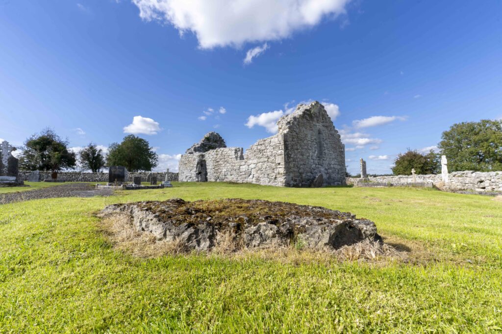 Ruins of an ancient stone building surrounded by green grass and old gravestones under a bright blue sky with scattered clouds.
